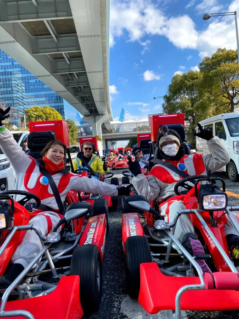 Group of people in red-and-white racing suits in go-karts on an outdoor track under an elevated road, waving at the camera.