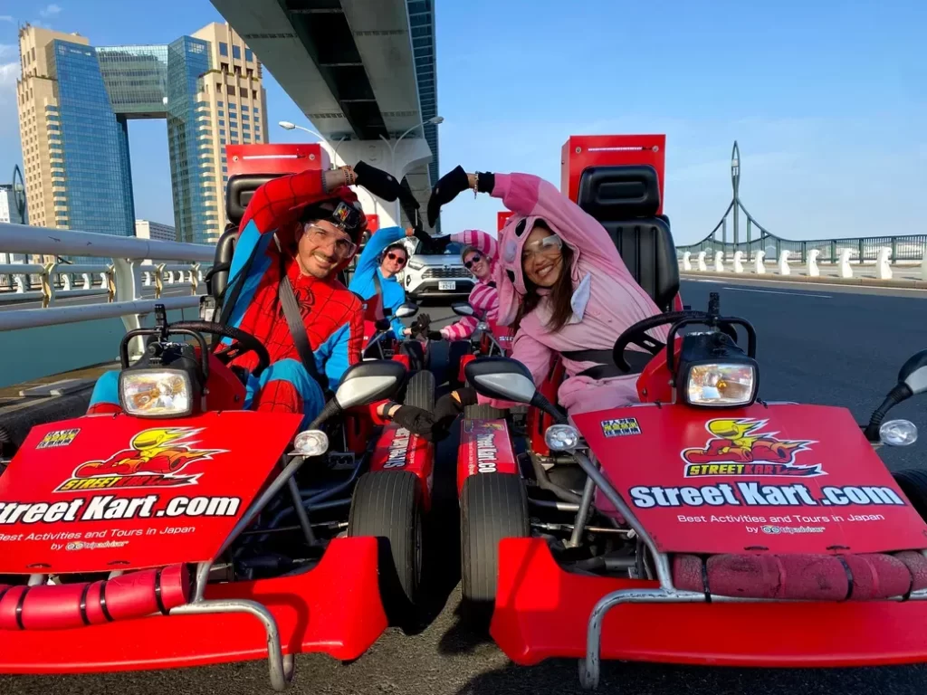 IMG_4112 - StreetKart Four people in red and pink go-karts on a sunny bridge, wearing colorful costumes (Spider-Man outfits) and forming a heart shape with their arms.