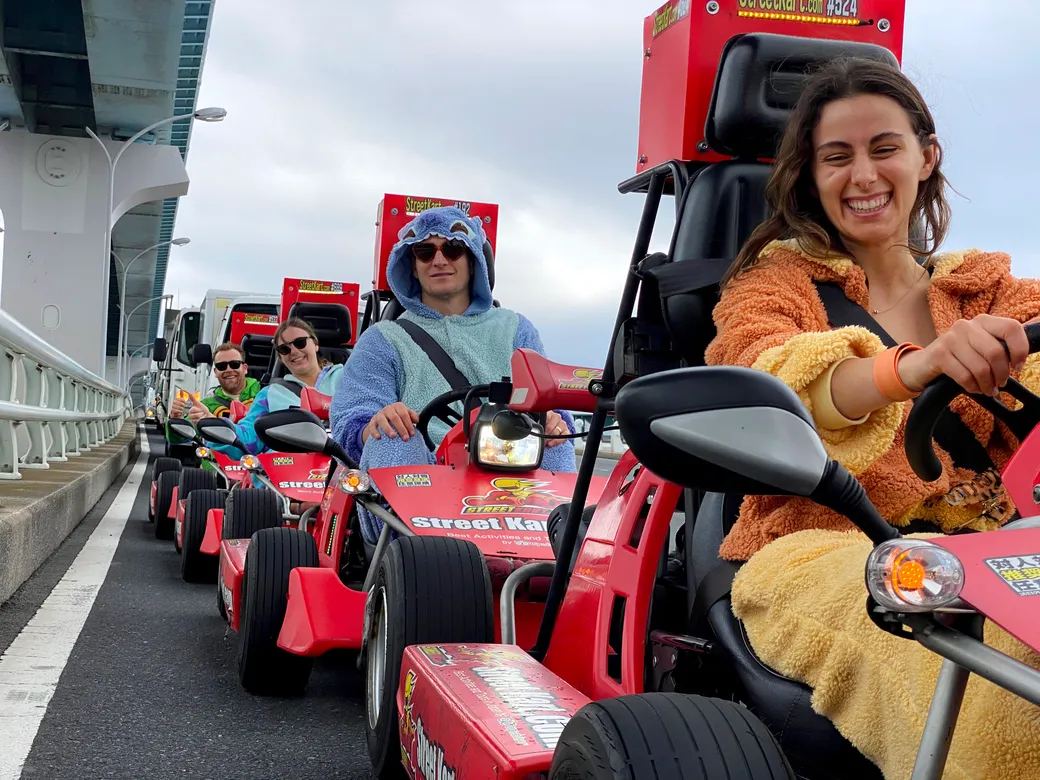 People in red go-karts wearing colorful costumes ride in a line on a road bridge, smiling and enjoying a group outing.